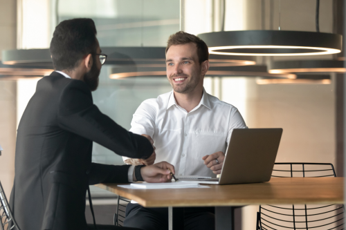 Aperto de mão entre dois profissionais numa sala de reuniões, com portátil sobre a mesa